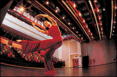 Dancer on stage mid-practice, bending towards an outstretched leg, the theatre seating is in the background